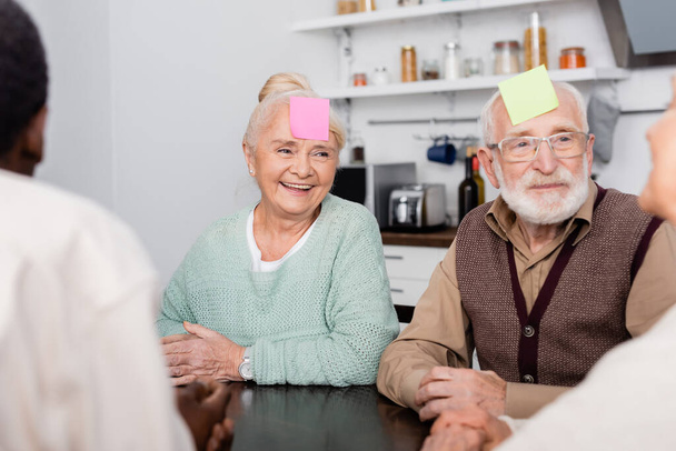 smiling senior man and woman with colorful sticky notes on foreheads playing game with multicultural friends on blurred foreground - Foto, Bild