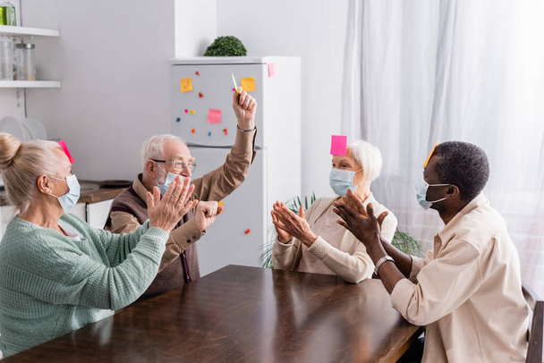 empolgados amigos seniores multiculturais em máscaras médicas com notas pegajosas na testa aplaudindo enquanto joga jogo na cozinha  - Foto, Imagem