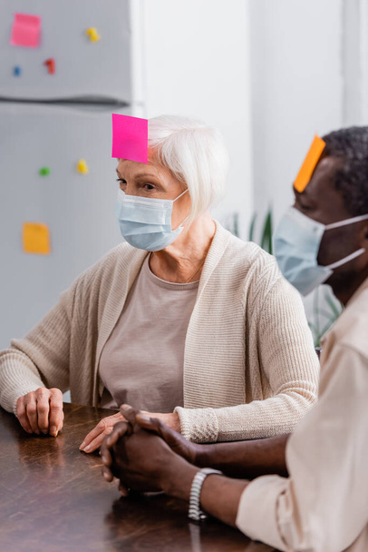 multicultural senior friends in medical masks with sticky notes on foreheads playing game in kitchen  - Fotografie, Obrázek