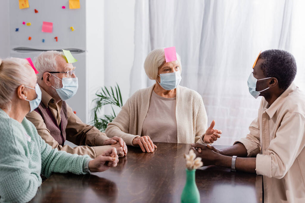 retraités portant des masques médicaux avec des notes collantes sur le front discutant tout en jouant au jeu avec des amis multiethniques - Photo, image