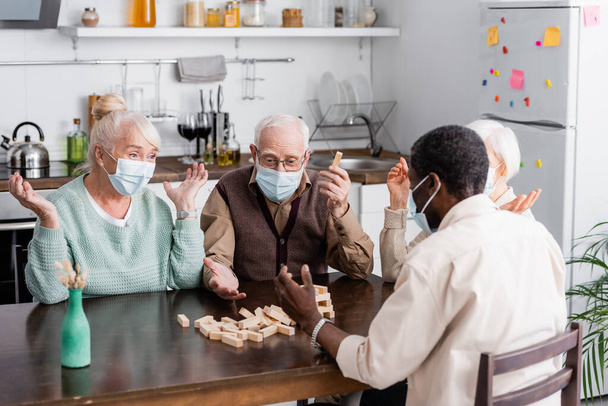 jubilados multiétnicos en máscaras médicas jugando torre bloques de madera juego en casa - Foto, Imagen