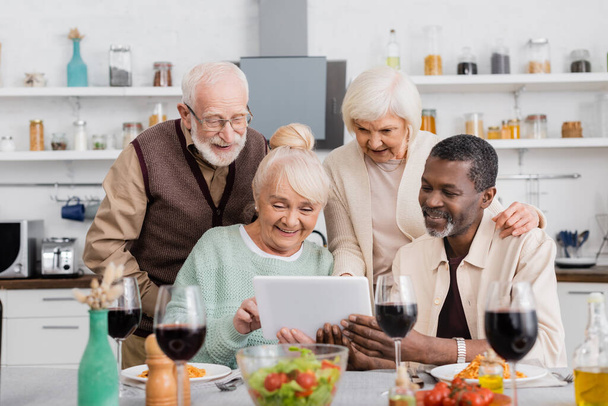 seniorów wielokulturowych osób patrząc na cyfrowy tablet ze szczęśliwymi przyjaciółmi w pobliżu smacznego jedzenia na stole - Zdjęcie, obraz