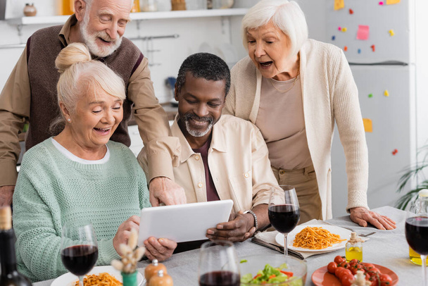 συνταξιούχοι πολυπολιτισμικοί άνθρωποι κοιτάζοντας ψηφιακή ταμπλέτα με χαρούμενους ηλικιωμένους φίλους κοντά νόστιμο φαγητό στο τραπέζι - Φωτογραφία, εικόνα
