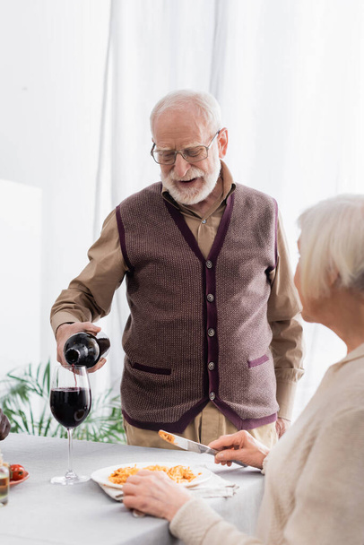 senior man in glasses pouring red wine in glass of retired woman - 写真・画像