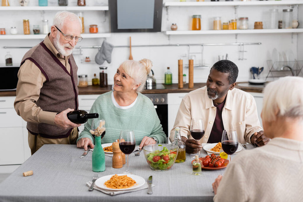 retired man in eyeglasses pouring red wine in glass near happy interracial friends  - Photo, Image