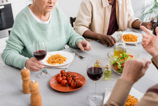 cropped view of senior woman holding glass of red wine while talking during lunch with multicultural friends on blurred background - Photo, Image