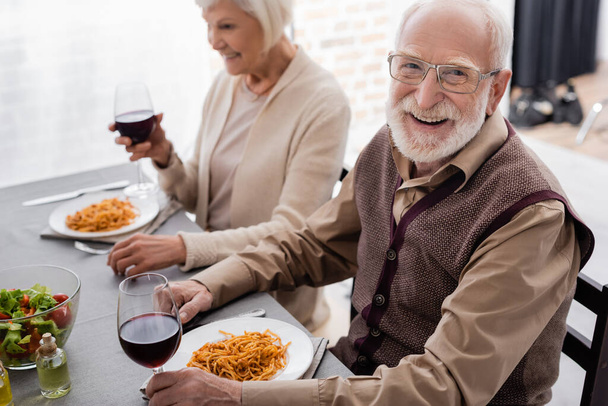 happy senior man in eyeglasses smiling while holding glass of wine during dinner with woman - Photo, Image