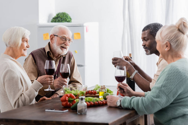 cheerful and retired interracial friends holding glasses with wine near vegetables on table - Photo, Image