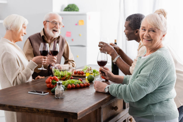 ευτυχισμένη ηλικιωμένη γυναίκα κρατώντας ένα ποτήρι κόκκινο κρασί και χαμογελώντας κοντά σε πολυπολιτισμικούς συνταξιούχους φίλους σε θολή φόντο - Φωτογραφία, εικόνα