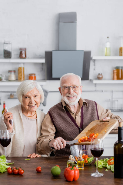 happy senior man adding sliced vegetables into bowl with salad near retired wife with smartphone  - Foto, Imagem