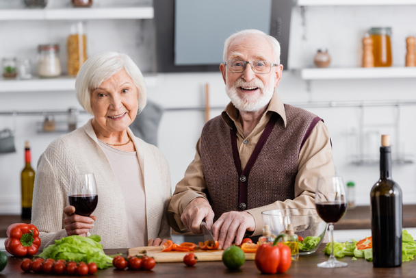 feliz hombre mayor cortando verduras cerca de la esposa jubilada con un vaso de vino - Foto, Imagen