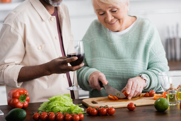 happy senior woman cutting ripe bell pepper near african american husband with glass of wine  - Foto, imagen