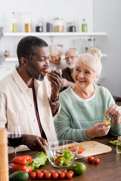 Uomo afroamericano mangiare pomodoro ciliegia vicino alla donna anziana e gli amici in pensione su sfondo sfocato  - Foto, immagini