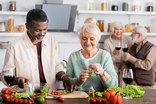 African american man and happy senior woman preparing salad near retired friends on blurred background  - Foto, Imagem