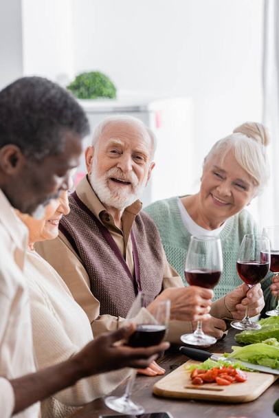 feliz jubilado sonriendo mientras sostiene una copa de vino cerca de amigos multiculturales en primer plano borroso - Foto, imagen
