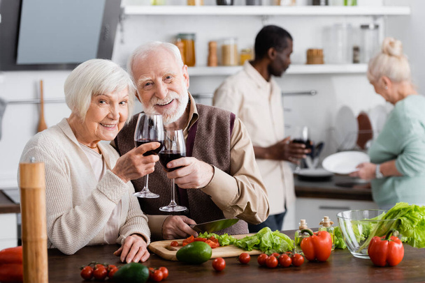 happy senior man and woman clinking glasses of wine near multicultural friends on blurred background  - Photo, Image
