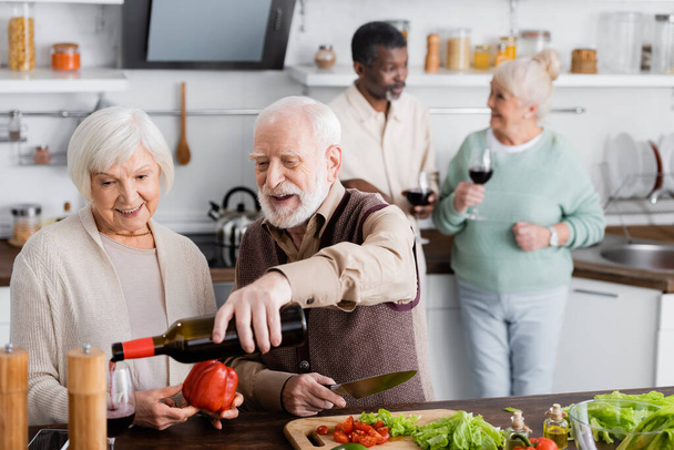 щасливий старший чоловік наливає вино в склянку біля пенсіонерів і мультикультурних друзів на розмитому фоні
  - Фото, зображення