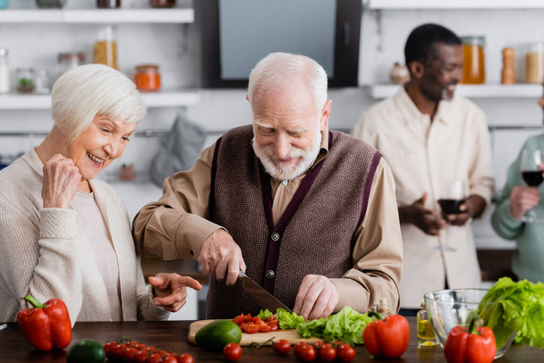 heureuse femme âgée pointant du doigt l'homme à la retraite cuisine salade près d'amis multiculturels sur fond flou  - Photo, image