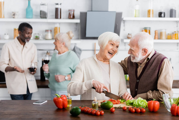 счастливый мужчина и женщина приготовления салата рядом с многонациональными друзьями на размытом фоне  - Фото, изображение