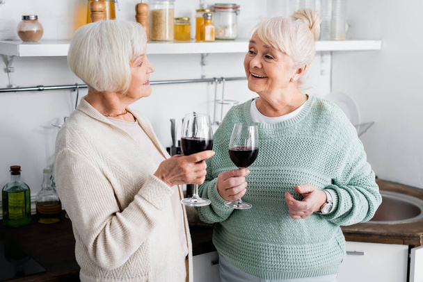 happy retired women holding glasses with wine and looking at each other in kitchen  - 写真・画像
