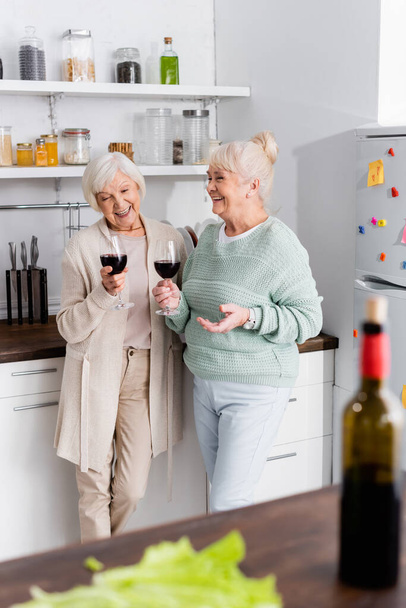 joyful retired women holding glasses with wine in kitchen  - 写真・画像
