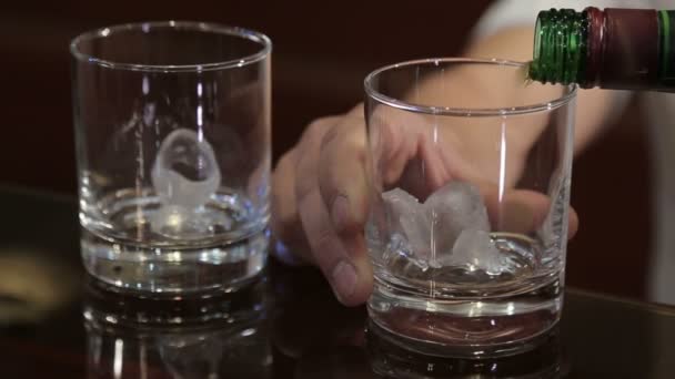 Barman versando whisky dorato, cognac o brandy dalla bottiglia in vetro con cubetti di ghiaccio sul tavolo di legno. Barista versando bicchieri di alcol bere bourbon, rum - Filmati, video