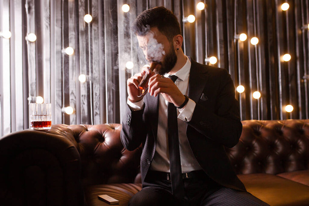 Portret brutalnego brodatego faceta z tatuażem palącego cygaro w eleganckim garniturze pijącego alkohol whisky w mieszkaniu na poddaszu. Koncepcja para love story - Zdjęcie, obraz