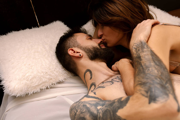 Beautiful loving couple Naked hug kissing in bed near the window in the morning . Romantic intimate lifestyle Love story of shirtless tattooed strong beard man with a sensual woman in loft apartment - Photo, Image