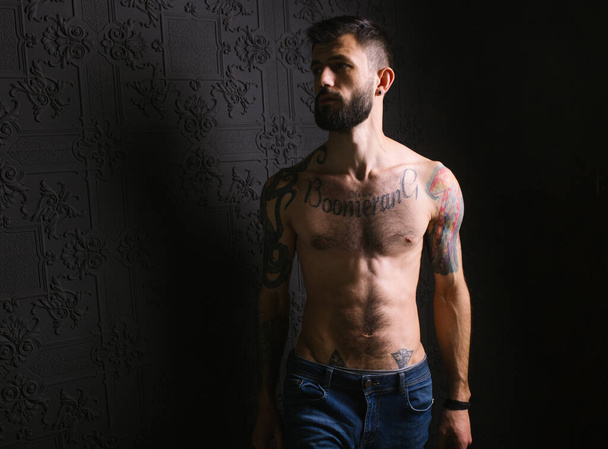 Portrét brutálního vousatého gentlemana s tetovaným tělem, kouřícího doutník v sexy elegantním obleku bez košile, nahého v podkrovním bytě. Silný sebevědomý potetovaný mužský kouř - Fotografie, Obrázek