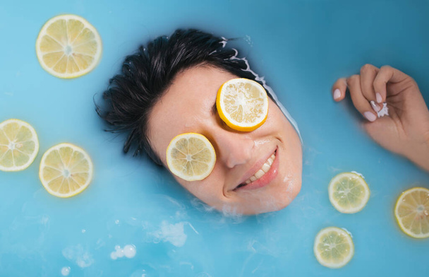 Attractive smiling girl takes a bath with milk . Spa treatments for skin rejuvenation . Cosmetology concept. Skin care product. Female portrait. Lemon slice face mask. Fruit scrub. Facial lifting - Photo, image