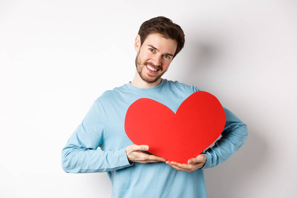 Charming lover man holding Valentines day red heart and smiling, I love you gesture, standing over white background - Zdjęcie, obraz
