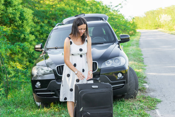  Νεαρή γυναίκα με ταξιδιωτική τσάντα στην άκρη του δρόμου κοντά στο αυτοκίνητο. Κορίτσι που έχει αυτοκίνητο περιπέτεια μεταφοράς. όμορφη κοπέλα σε λευκό φόρεμα με αποσκευές - Φωτογραφία, εικόνα