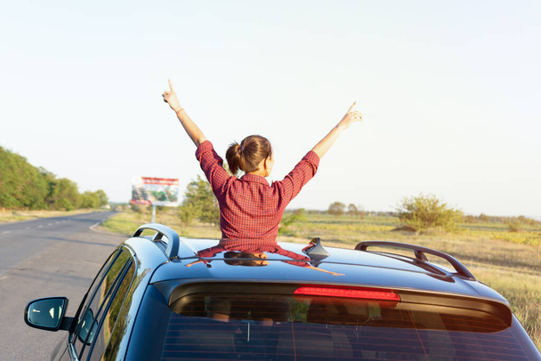 Ταξιδεύοντας με το αυτοκίνητο. Το κορίτσι κοιτάζει έξω από την καταπακτή του αυτοκινήτου. νεαρή γυναίκα κάθεται στην οροφή του αυτοκινήτου και να απολαύσετε τη ζωή - Φωτογραφία, εικόνα
