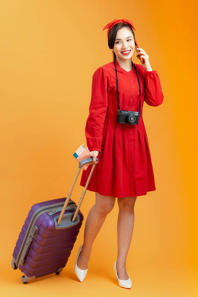 Happy Asian woman holding suitcase and passport with flight tickets while talking on phone, isolated on orange background - Фото, изображение
