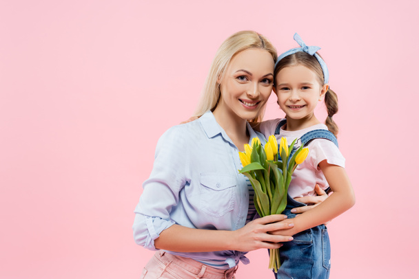 happy mother holding yellow tulips and hugging daughter isolated on pink  - Foto, immagini