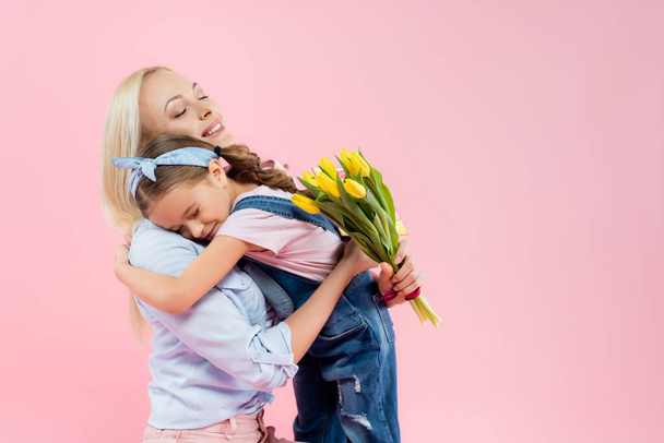 cheerful mother holding yellow tulips and hugging daughter isolated on pink  - Foto, Imagem