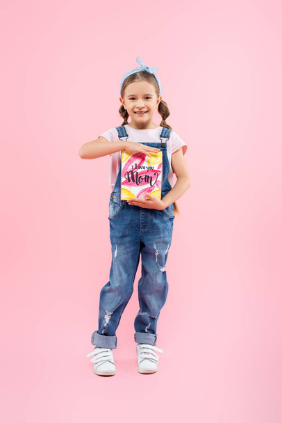 full length of happy kid holding greeting card with i love you mom lettering on pink  - Photo, Image