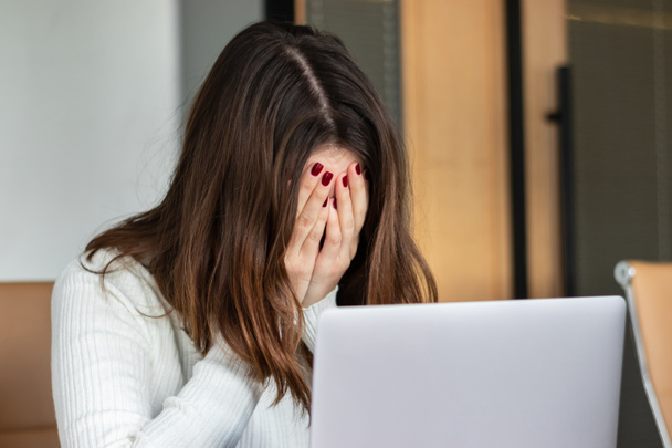 Θλιβερή και κουρασμένη γυναίκα με λευκό πουλόβερ που εργάζεται με φορητό υπολογιστή στο γραφείο - Φωτογραφία, εικόνα