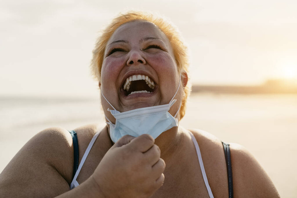 Szczęśliwy plus rozmiar kobieta śmiejąc się na plaży podczas noszenia maski twarzy - Curvy model z nadwagą bawiąc się podczas wakacji w tropikalnym miejscu - Opieka zdrowotna i ponadgabarytowa pewność koncepcja osoby - Zdjęcie, obraz