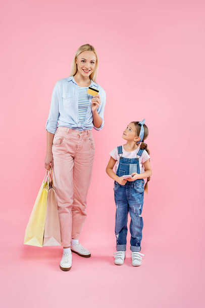 full length of mother holding credit card and shopping bags near daughter with smartphone on pink  - Fotoğraf, Görsel