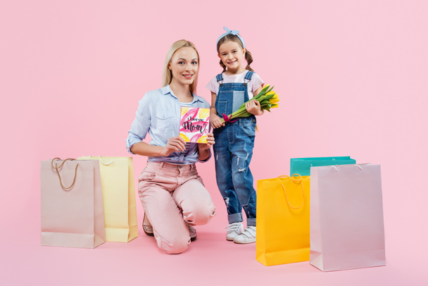 full length of mother holding greeting card near kid with flowers and shopping bags on pink - Photo, Image