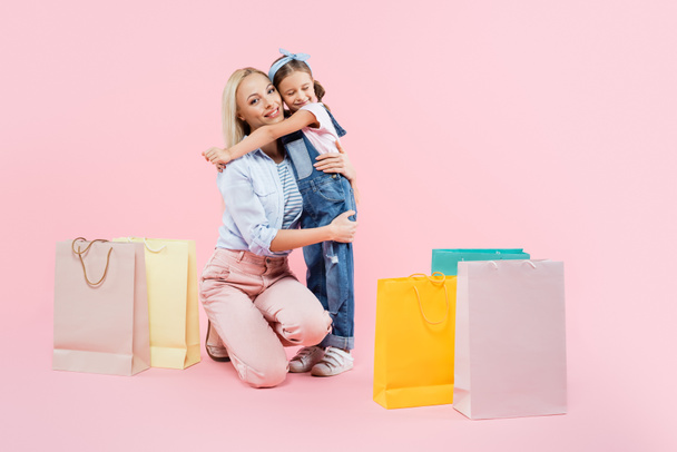 full length of happy daughter hugging smiling mother near shopping bags on pink  - Foto, afbeelding