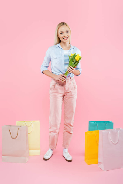 full length of happy woman holding yellow tulips near shopping bags on pink - Foto, Bild