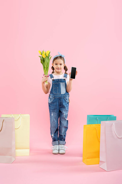 full length of kid holding smartphone with blank screen and tulips near shopping bags on pink - Фото, зображення