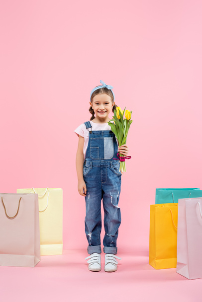 full length of happy kid holding tulips near shopping bags on pink - Φωτογραφία, εικόνα