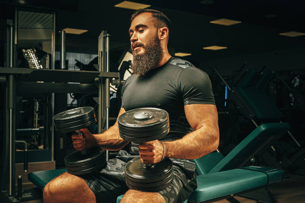 Muscular young man lifting weights in a dark gym - Zdjęcie, obraz