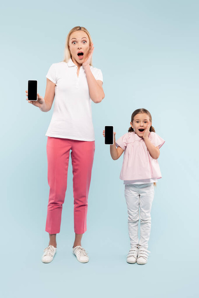 full length of shocked mother and daughter holding smartphones with blank screen on blue - Foto, Bild