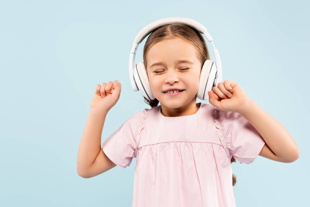 happy kid with closed eyes in wireless headphones smiling isolated on blue - Φωτογραφία, εικόνα