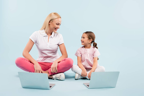 happy mother and daughter sitting with crossed legs near laptops and looking at each other on blue - Foto, Imagem
