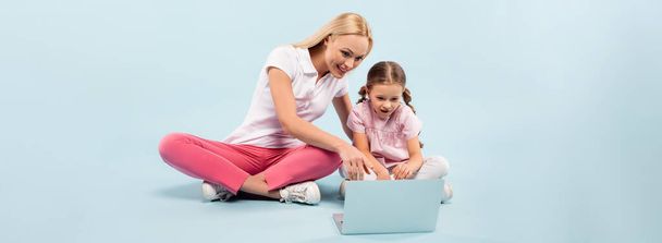 smiling mother and daughter sitting with crossed legs and pointing with finger laptop on blue, banner - Foto, Imagen